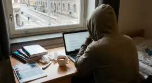 An international student in a hoodie sits at a desk in a Gothenburg dorm, looking stressed and unwell while searching for healthcare on a laptop. The desk is cluttered with tissues, a thermometer, a cup of tea, and an Erasmus brochure.
